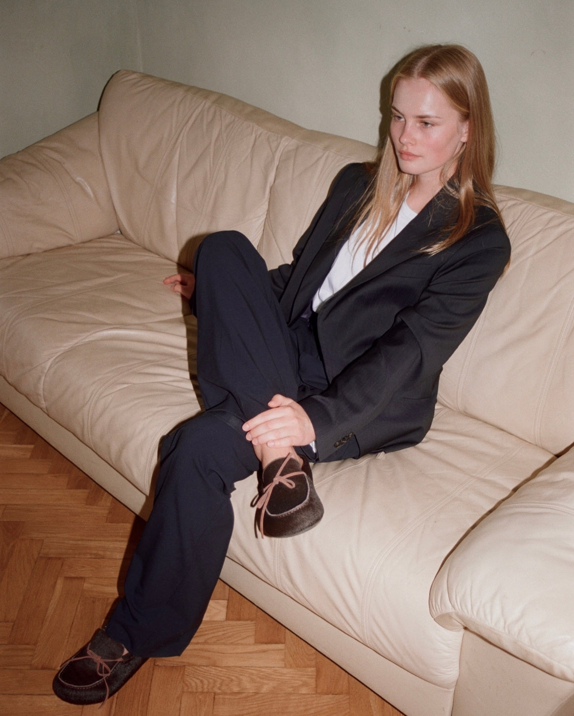 A woman sits on a cream-colored leather sofa wearing a black blazer and trousers with brown lace-up clog slippers over a white shirt.