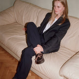 A woman sits on a cream-colored leather sofa wearing a black blazer and trousers with brown lace-up clog slippers over a white shirt.
