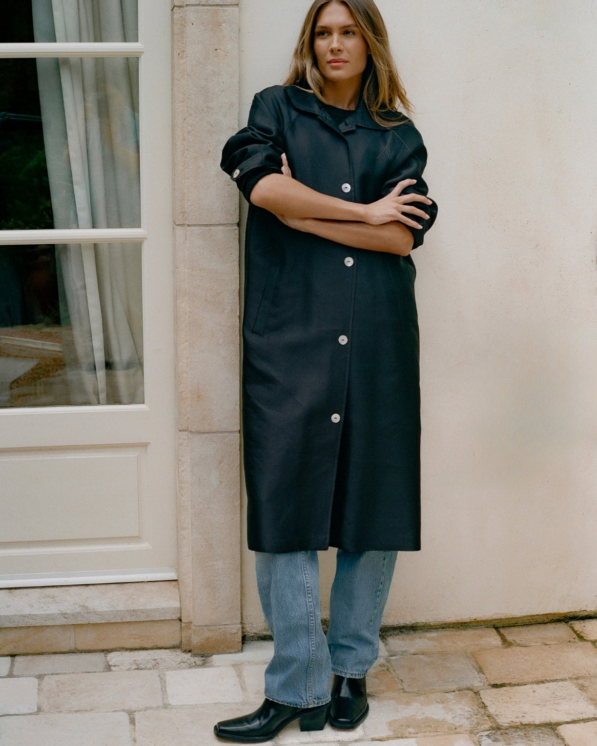 A woman stands outdoors against a beige wall and stone column, wearing a long black coat with silver-tone buttons, light blue jeans, and black ankle boots.