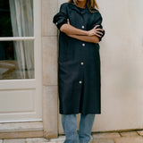 A woman stands outdoors against a beige wall and stone column, wearing a long black coat with silver-tone buttons, light blue jeans, and black ankle boots.