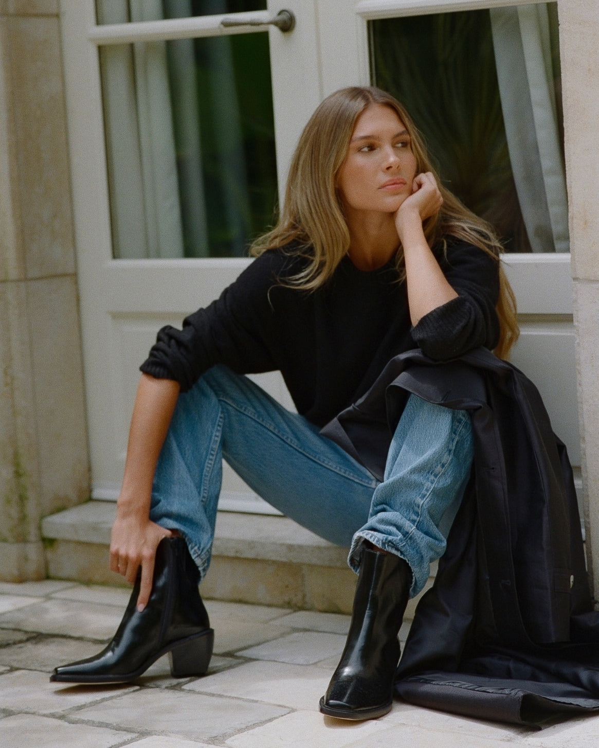 A woman sits on stone steps outside a building, wearing black ankle boots, a black long-sleeve top, and blue jeans, with a black coat draped over her leg.