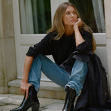 A woman sits on stone steps outside a building, wearing black ankle boots, a black long-sleeve top, and blue jeans, with a black coat draped over her leg.