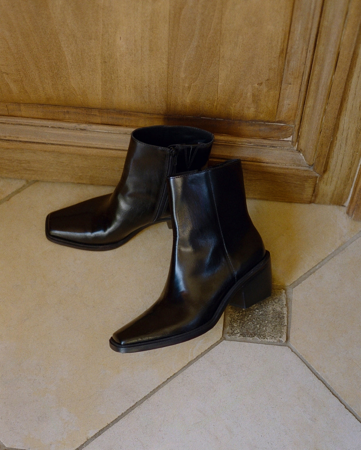 A pair of black leather ankle boots with a block heel and pointed toe, resting on a beige tiled floor in front of a wooden door.