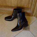 A pair of black leather ankle boots with a block heel and pointed toe, resting on a beige tiled floor in front of a wooden door.