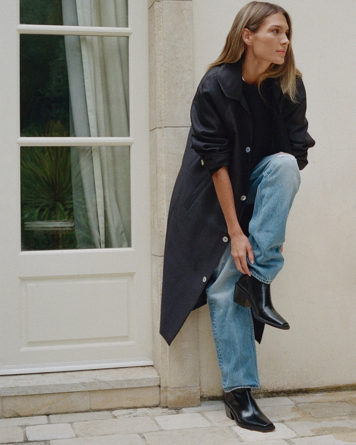 A woman models a long black trench coat over a black top and light blue jeans, leaning against a beige stone wall next to a white paneled door.
