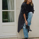 A woman models a long black trench coat over a black top and light blue jeans, leaning against a beige stone wall next to a white paneled door.