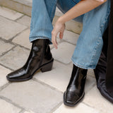 A person wearing shiny black ankle boots with a low block heel and square toes, paired with light blue jeans, sitting on a stone patio.
