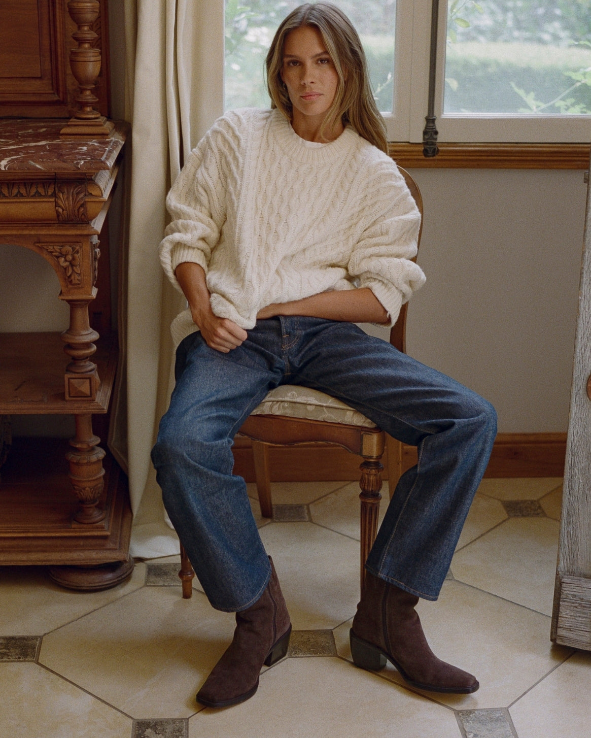 A woman sits on a wooden chair in a rustic room, wearing a cream cable-knit sweater, blue jeans, and dark brown ankle boots.