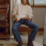 A woman sits on a wooden chair in a rustic room, wearing a cream cable-knit sweater, blue jeans, and dark brown ankle boots.