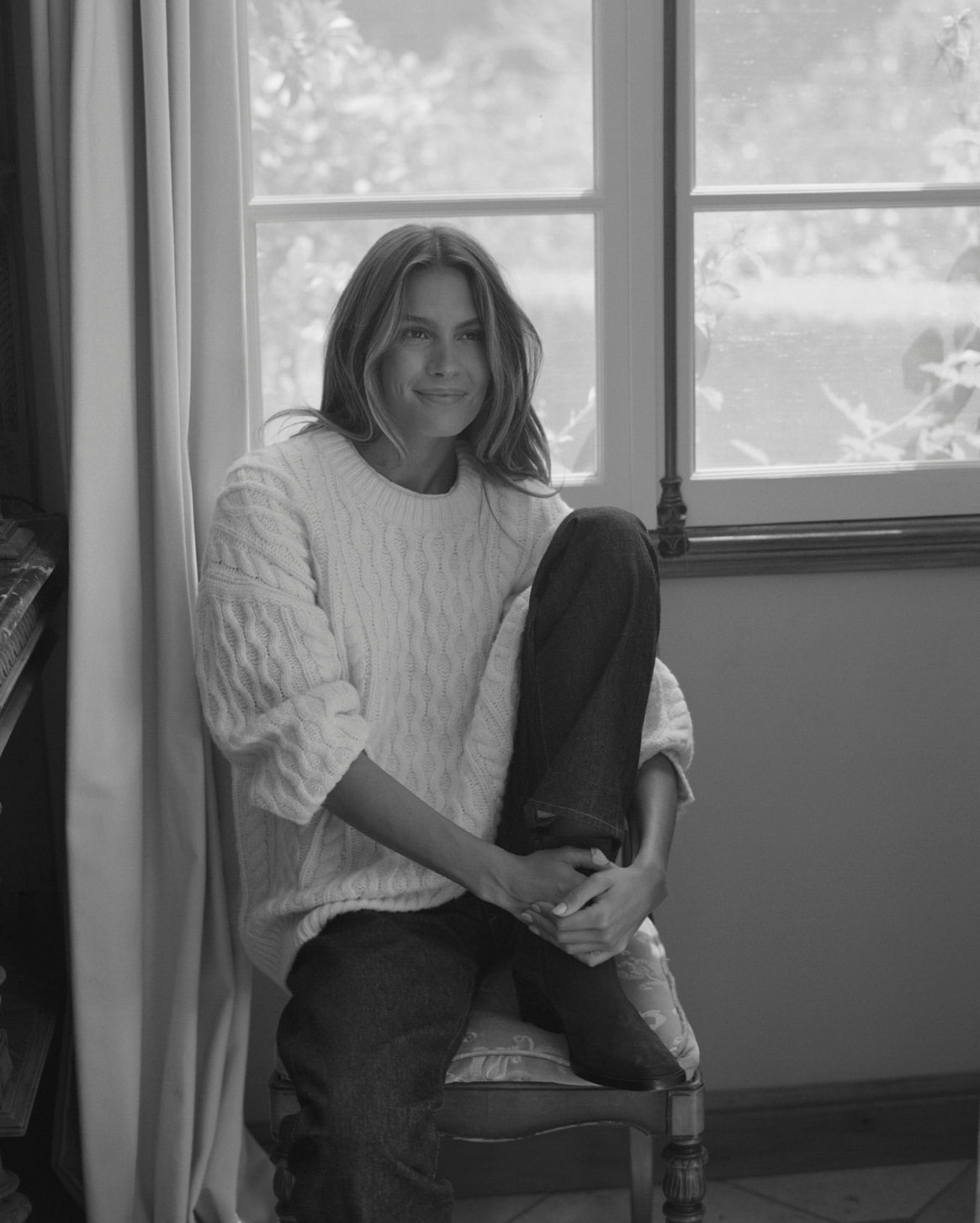 A black and white portrait of a woman sitting on a chair in front of a window, wearing a textured cable-knit sweater and dark pants.