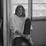 A black and white portrait of a woman sitting on a chair in front of a window, wearing a textured cable-knit sweater and dark pants.