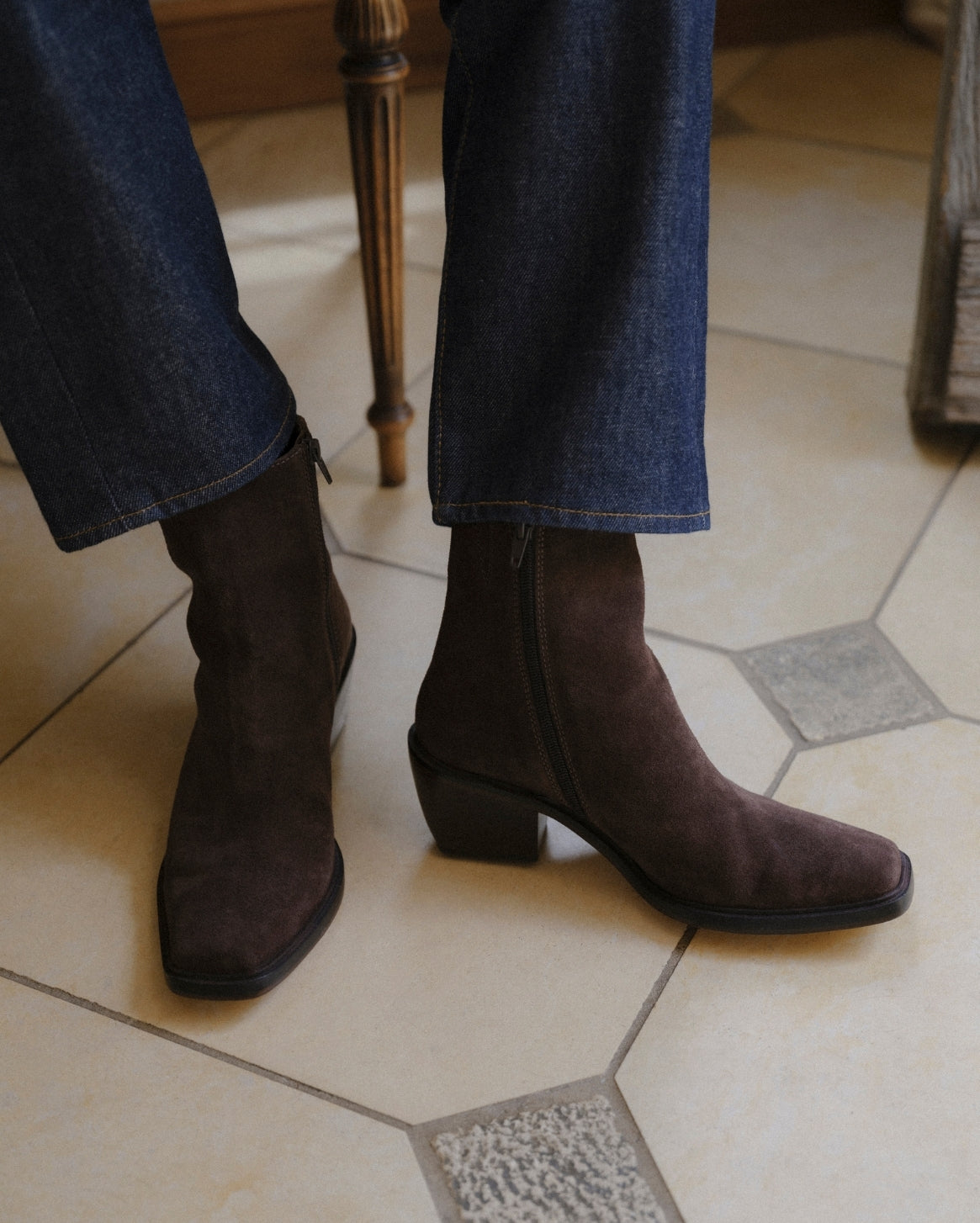 A person wearing dark brown suede ankle boots with low block heels and square toes, paired with blue jeans, standing on a beige tiled floor.