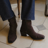 A person wearing dark brown suede ankle boots with low block heels and square toes, paired with blue jeans, standing on a beige tiled floor.