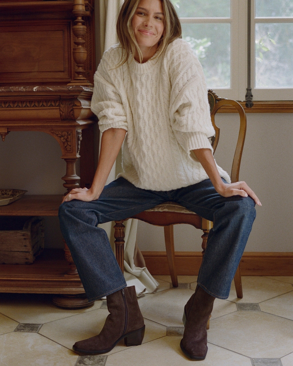 A woman sits on a wooden chair in a rustic room, wearing a chunky cream cable-knit sweater, dark blue jeans, and brown ankle boots.