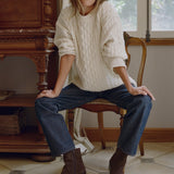 A woman sits on a wooden chair in a rustic room, wearing a chunky cream cable-knit sweater, dark blue jeans, and brown ankle boots.