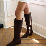 A person wearing dark brown suede knee-high boots with low block heels, standing on a wooden floor in a bright room with white walls and a window.