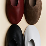 Four minimalist slip-on flats arranged in a 2x2 grid on a light beige background, showing different color options: burgundy leather, brown suede, black leather, and white leather.
