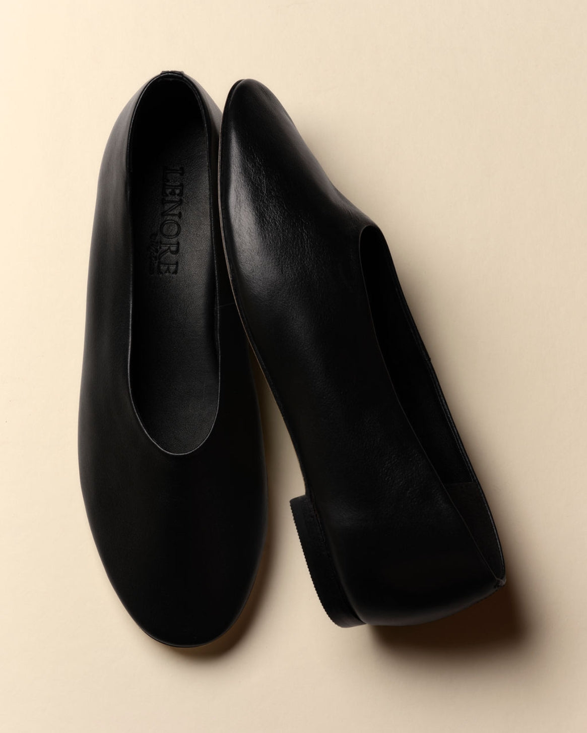 A pair of black leather slip-on flats with a low block heel, shown from top and side angles on a warm beige background. The brand name 'LENORE' is debossed on the insole of the left shoe.