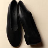 A pair of black leather slip-on flats with a low block heel, shown from top and side angles on a warm beige background. The brand name 'LENORE' is debossed on the insole of the left shoe.