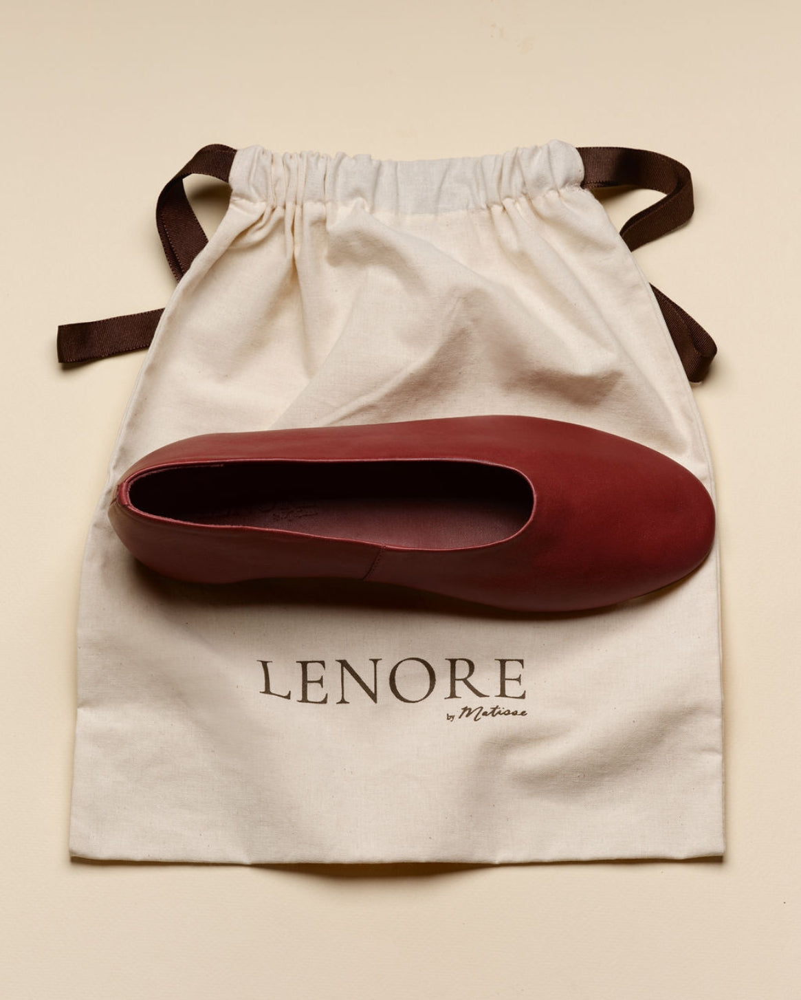 A burgundy leather ballet flat shoe resting on a cream-colored dust bag with the brand name 'LENORE by Matisse' printed on it, set against a solid beige background.