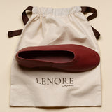 A burgundy leather ballet flat shoe resting on a cream-colored dust bag with the brand name 'LENORE by Matisse' printed on it, set against a solid beige background.