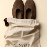 A pair of dark brown suede slip-on shoes resting on top of a cream-colored dust bag with the brand name 'LENORE' printed on it, set against a solid light beige background.
