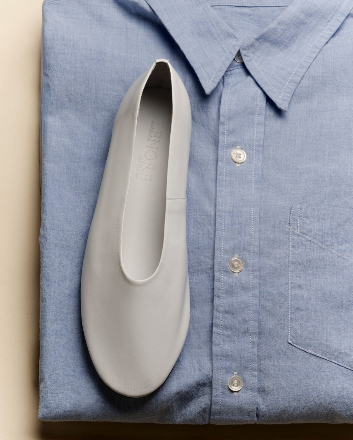A white slip-on shoe placed on top of a folded light blue button-up shirt, photographed from above against a warm beige background.