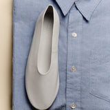 A white slip-on shoe placed on top of a folded light blue button-up shirt, photographed from above against a warm beige background.
