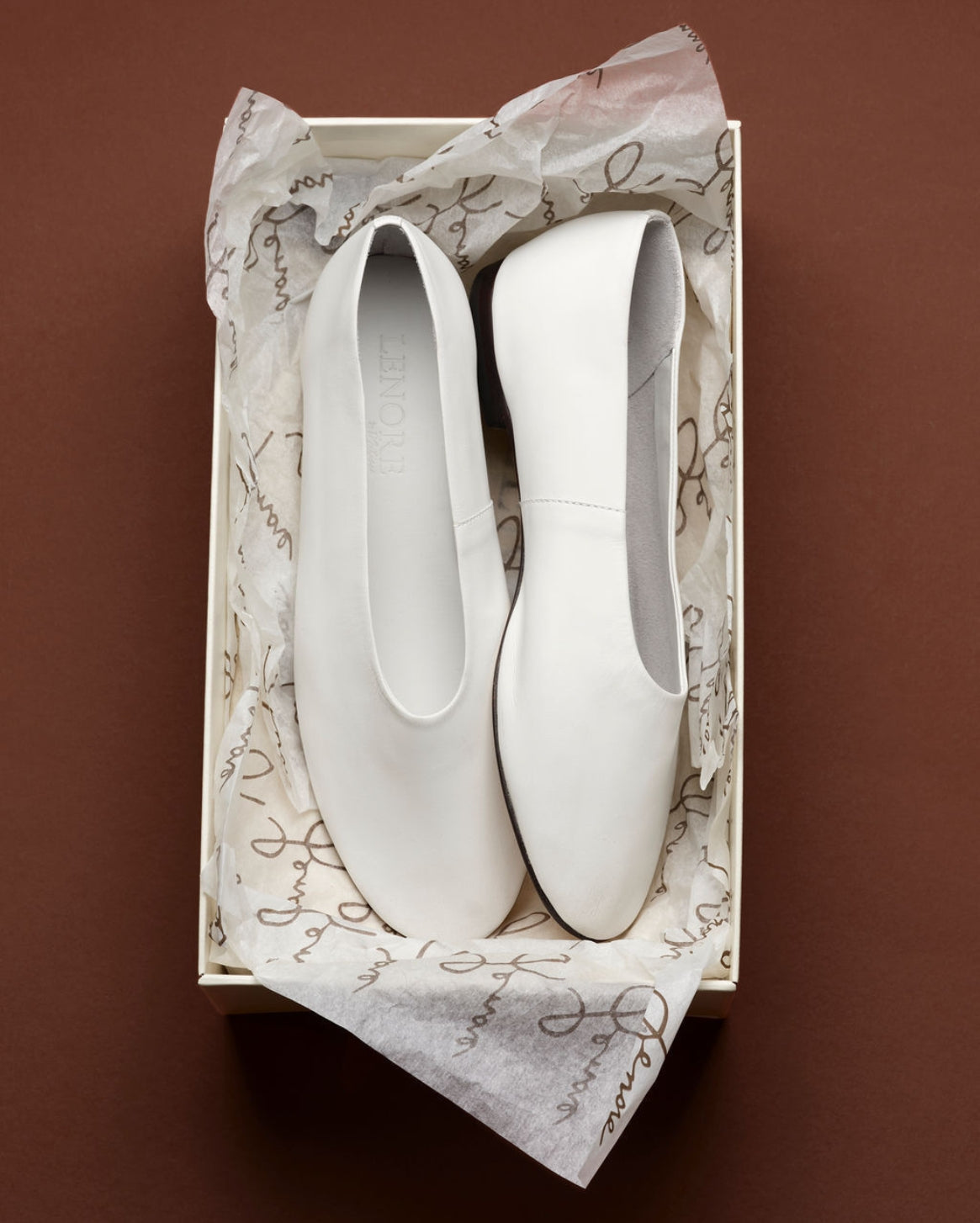 A pair of white leather slip-on flats resting inside a cream-colored shoebox lined with white patterned tissue paper, set against a solid dark brown background.