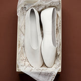 A pair of white leather slip-on flats resting inside a cream-colored shoebox lined with white patterned tissue paper, set against a solid dark brown background.