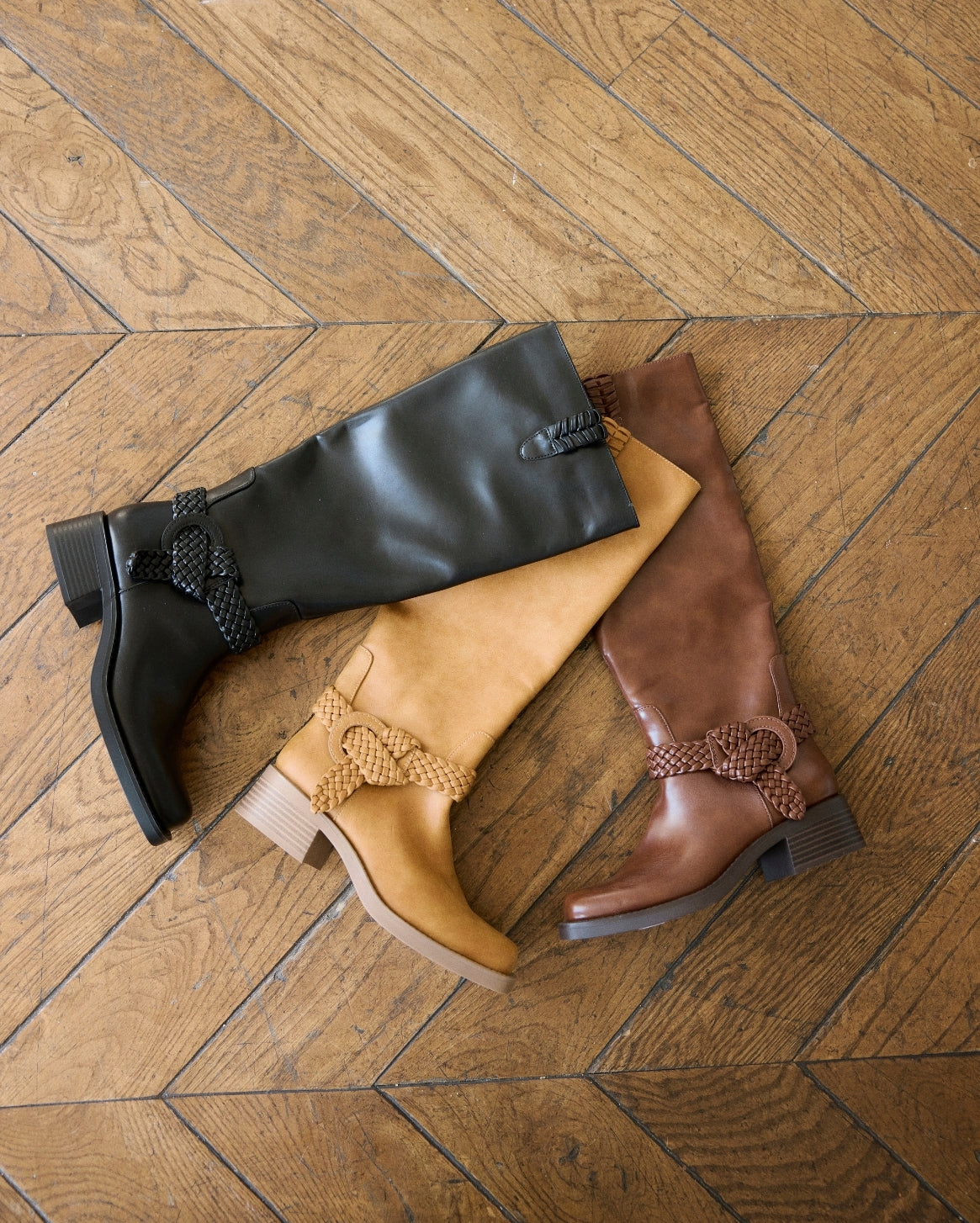 Three knee-high leather boots in black, tan, and brown, arranged on a wooden herringbone floor. Each boot features a braided decorative detail near the ankle and a low block heel.