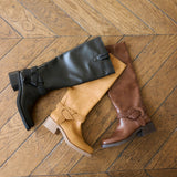 Three knee-high leather boots in black, tan, and brown, arranged on a wooden herringbone floor. Each boot features a braided decorative detail near the ankle and a low block heel.