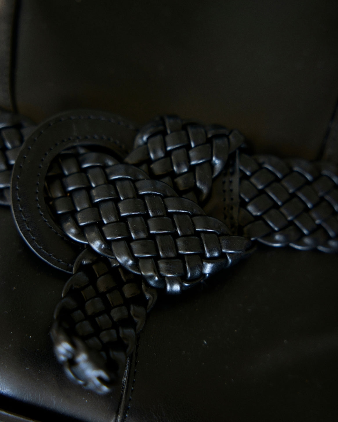 A close-up of a black braided leather belt with a large round metal ring buckle, resting on a dark leather surface.