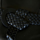 A close-up of a black braided leather belt with a large round metal ring buckle, resting on a dark leather surface.