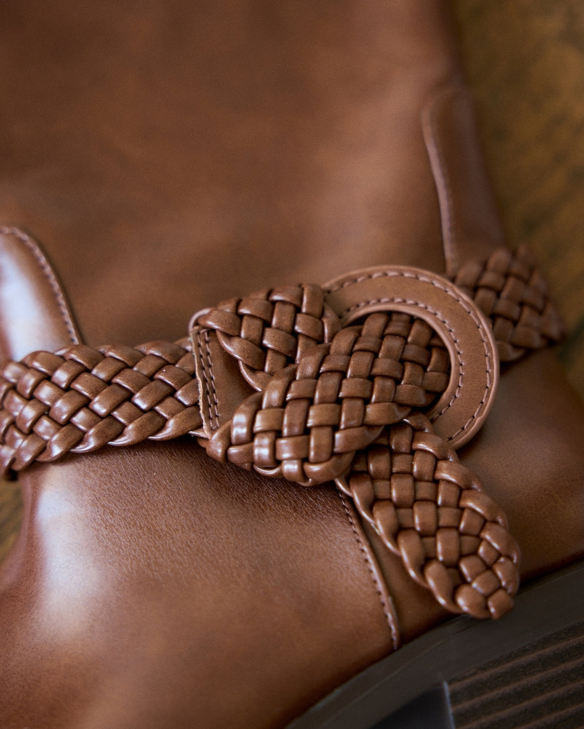 A close-up of a brown leather boot featuring a decorative braided leather strap tied in a bow around the ankle.