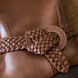 A close-up of a brown leather boot featuring a decorative braided leather strap tied in a bow around the ankle.