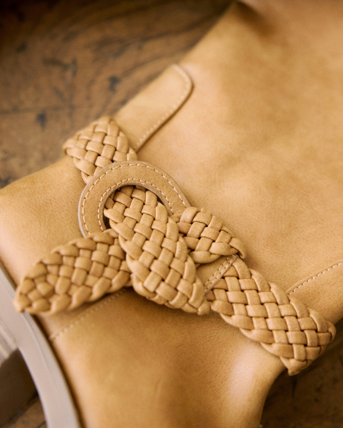 A close-up of a tan leather boot featuring a decorative braided leather strap tied in a bow around the ankle.