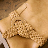 A close-up of a tan leather boot featuring a decorative braided leather strap tied in a bow around the ankle.