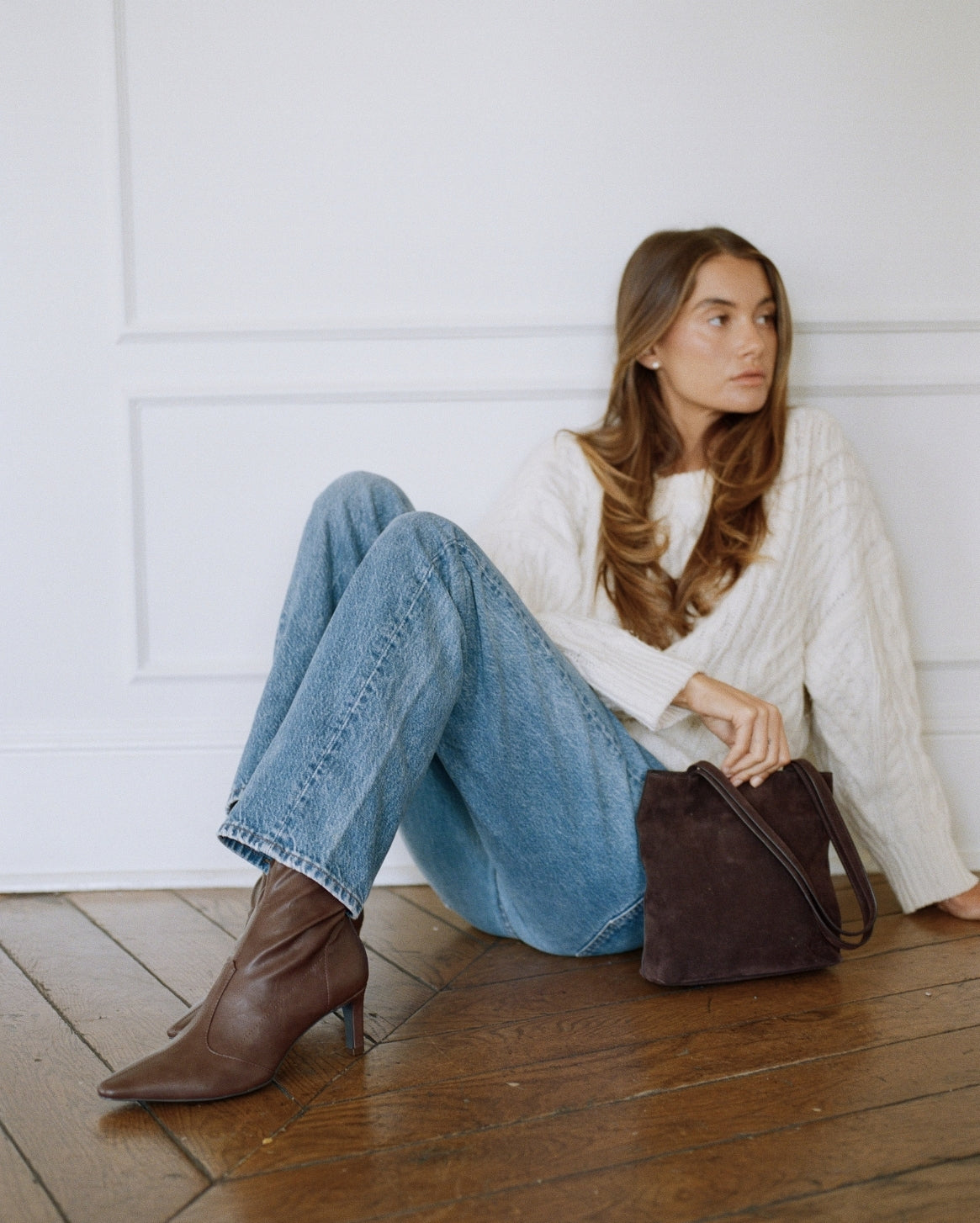 A woman sits on a wooden floor against a white paneled wall, wearing a white cable-knit sweater, blue jeans, and brown ankle boots, with a dark brown suede shoulder bag beside her.