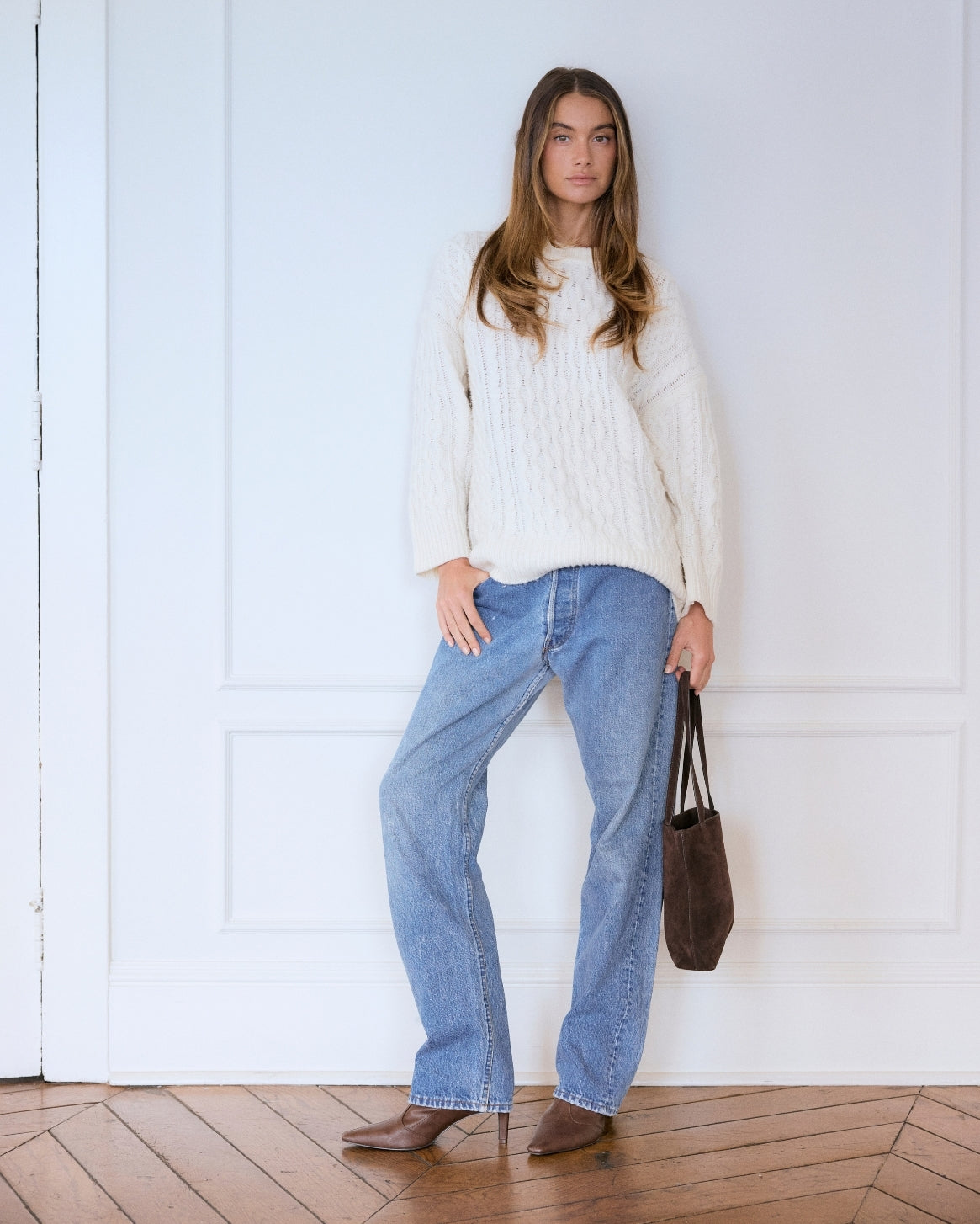 A woman stands against a white paneled wall wearing a cream cable-knit sweater, wide-leg light blue jeans, and brown ankle boots, while holding a dark brown tote bag.