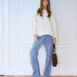 A woman stands against a white paneled wall wearing a cream cable-knit sweater, wide-leg light blue jeans, and brown ankle boots, while holding a dark brown tote bag.