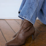 A pair of brown leather pointed-toe ankle boots with kitten heels, shown on a model wearing blue jeans on a wooden floor.
