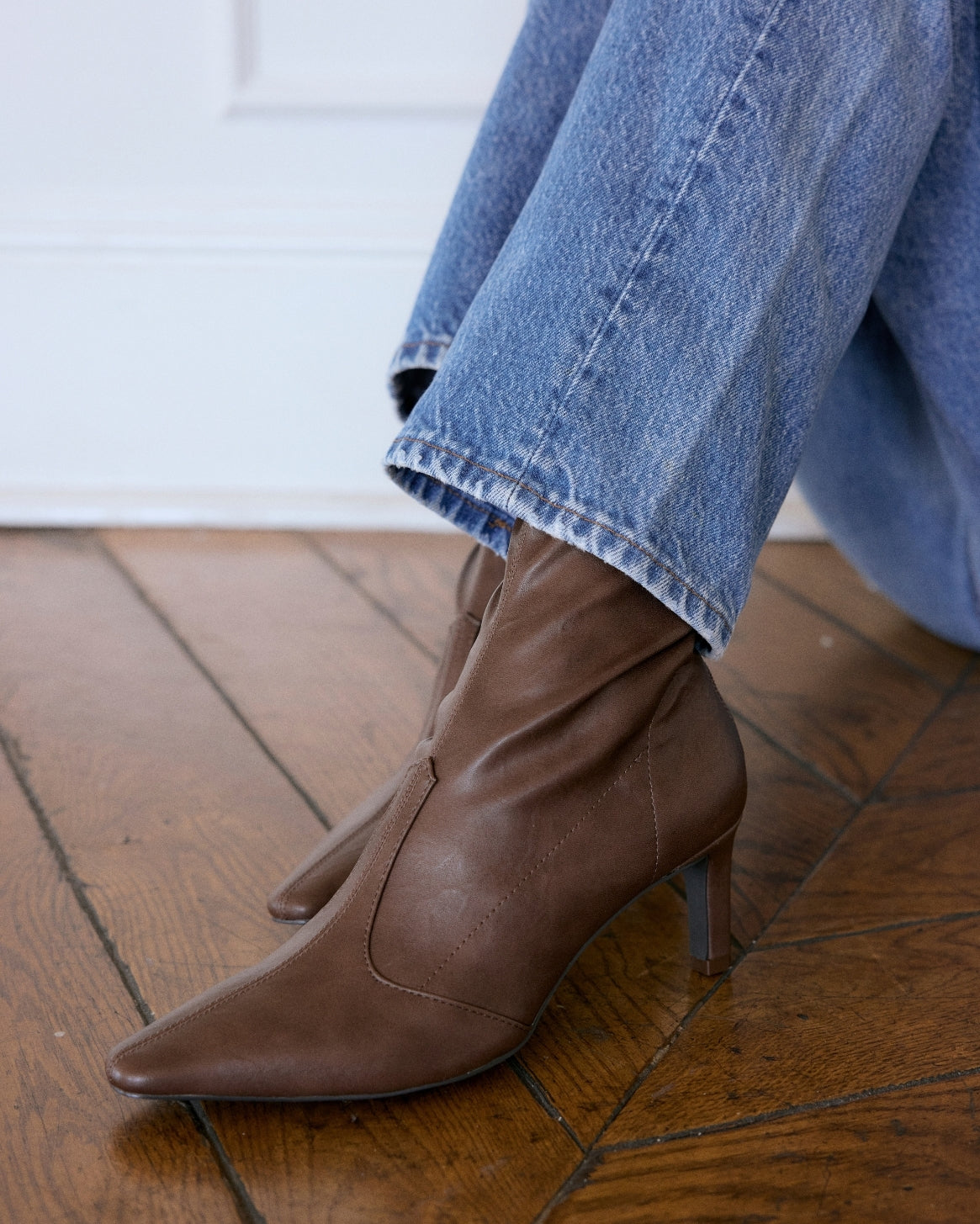 A pair of brown leather pointed-toe ankle boots with kitten heels, shown on a model wearing blue jeans on a wooden floor.