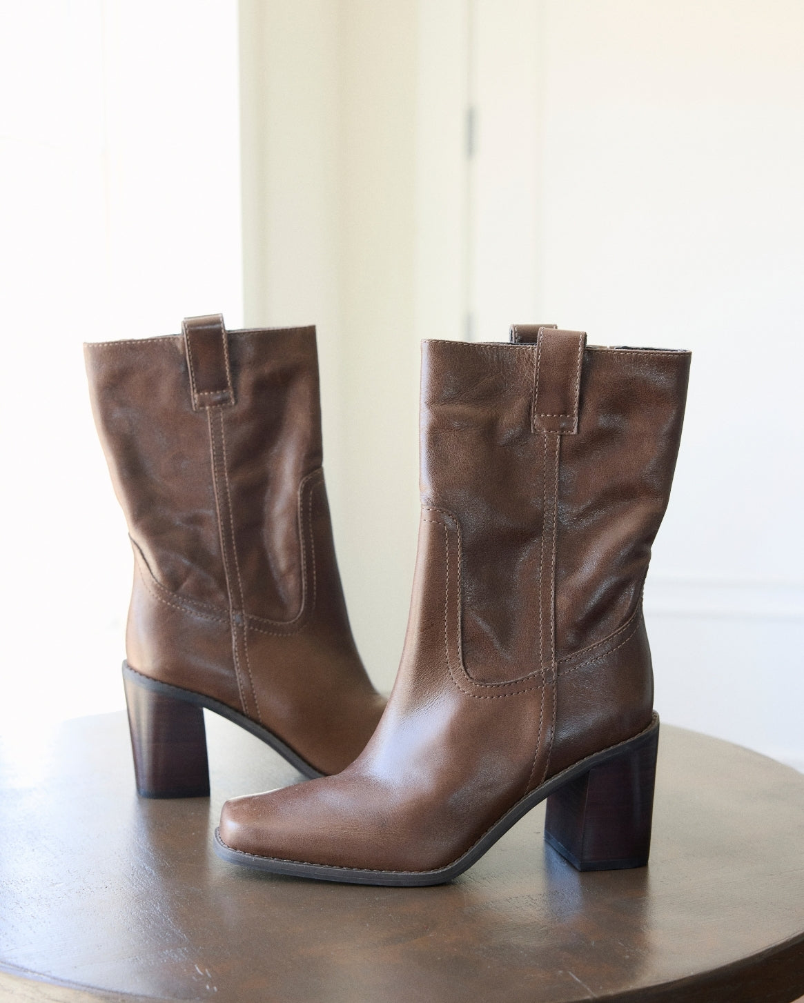 A pair of brown leather mid-calf boots with block heels, displayed on a dark wooden table against a bright, softly lit indoor background.