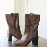 A pair of brown leather mid-calf boots with block heels, displayed on a dark wooden table against a bright, softly lit indoor background.