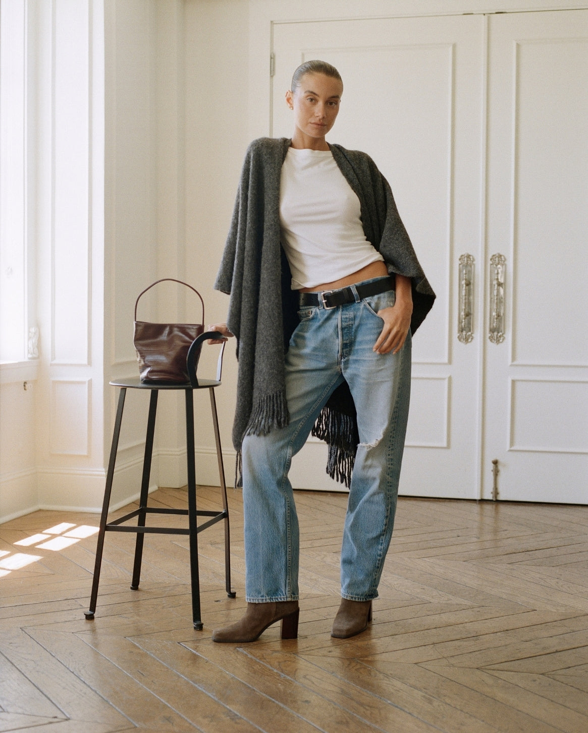 A woman stands in a bright room with white paneled doors and wood floors, wearing a white top, light blue jeans, a dark gray fringed cardigan, and brown ankle boots. A brown handbag sits on a black stool beside her.