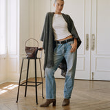 A woman stands in a bright room with white paneled doors and wood floors, wearing a white top, light blue jeans, a dark gray fringed cardigan, and brown ankle boots. A brown handbag sits on a black stool beside her.