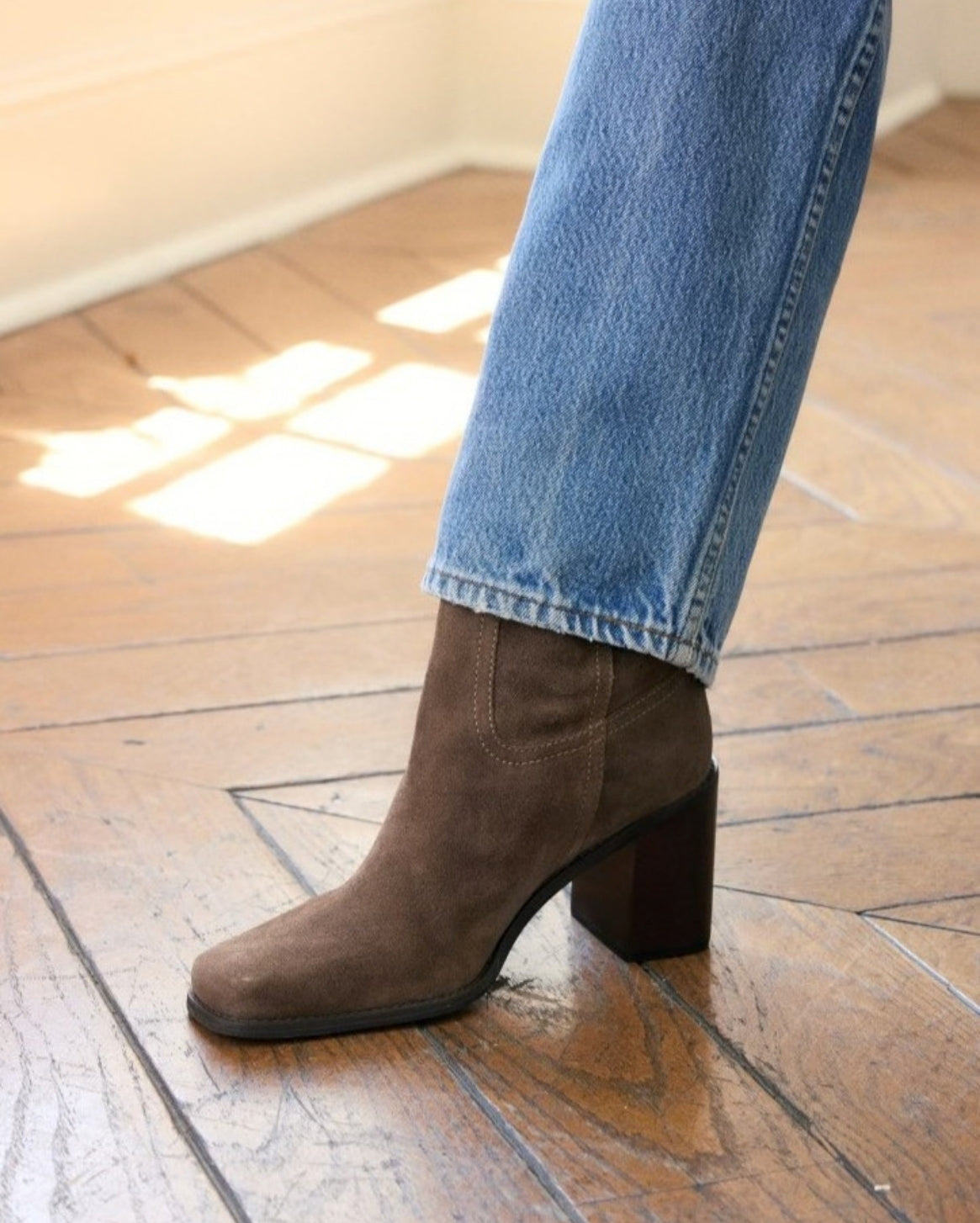 A person wearing a brown suede ankle boot with a block heel and cuffed blue jeans, standing on a wooden floor.