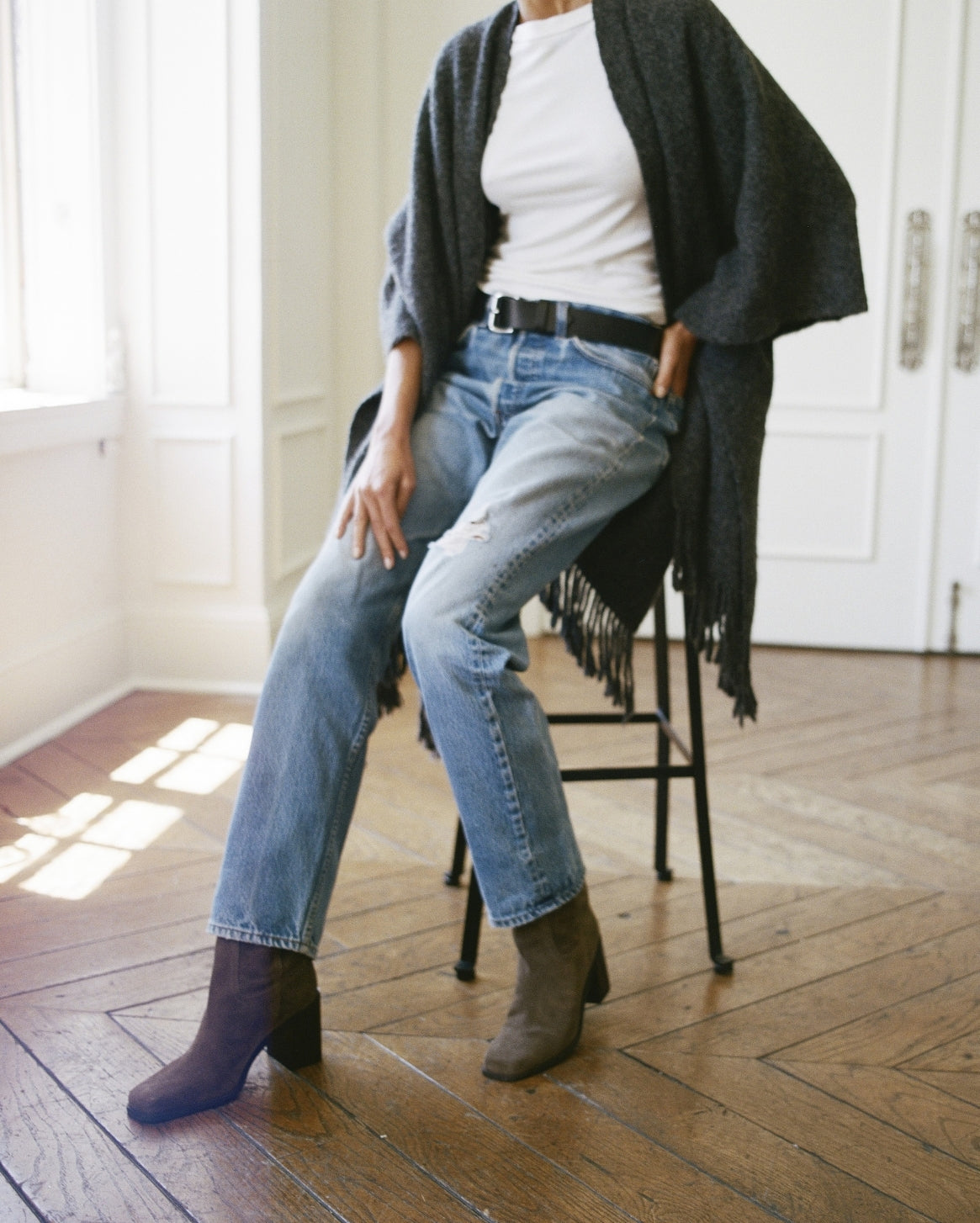 A person seated on a black stool in a bright room with wood floors, wearing light blue straight-leg jeans, a white top, a dark charcoal fringed cardigan, and dark brown ankle boots.