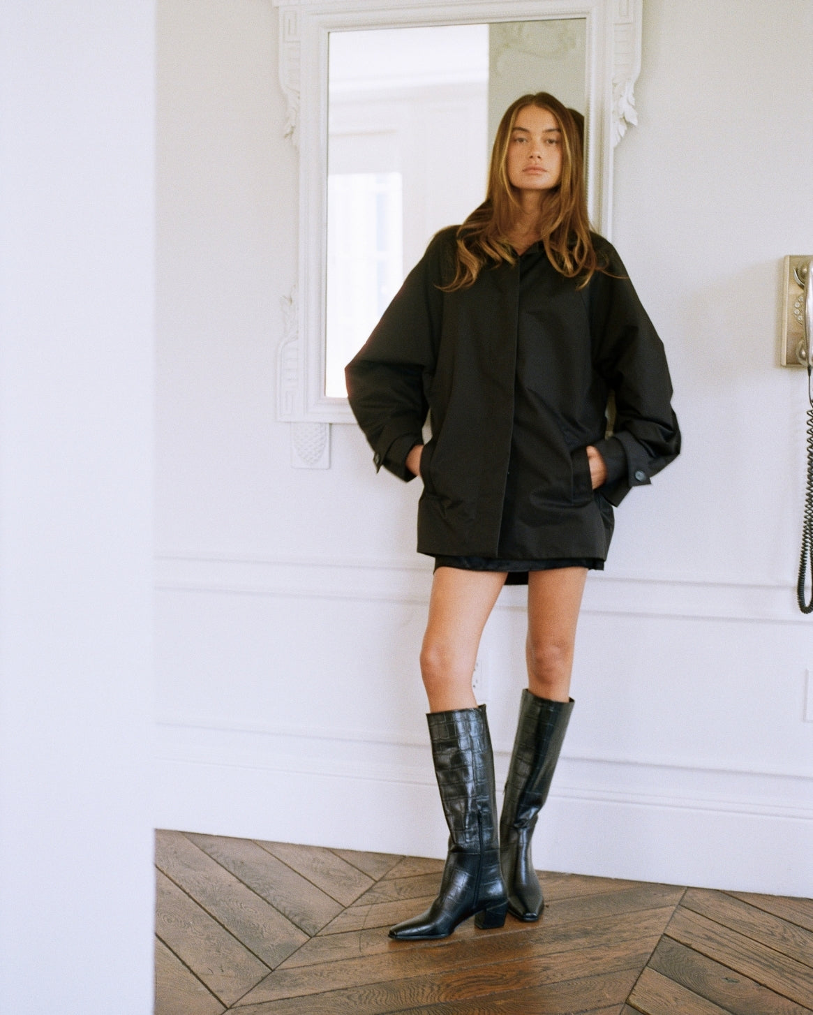 A woman stands in a bright white room with a wood herringbone floor, wearing an oversized black coat, black shorts, and black knee-high boots.